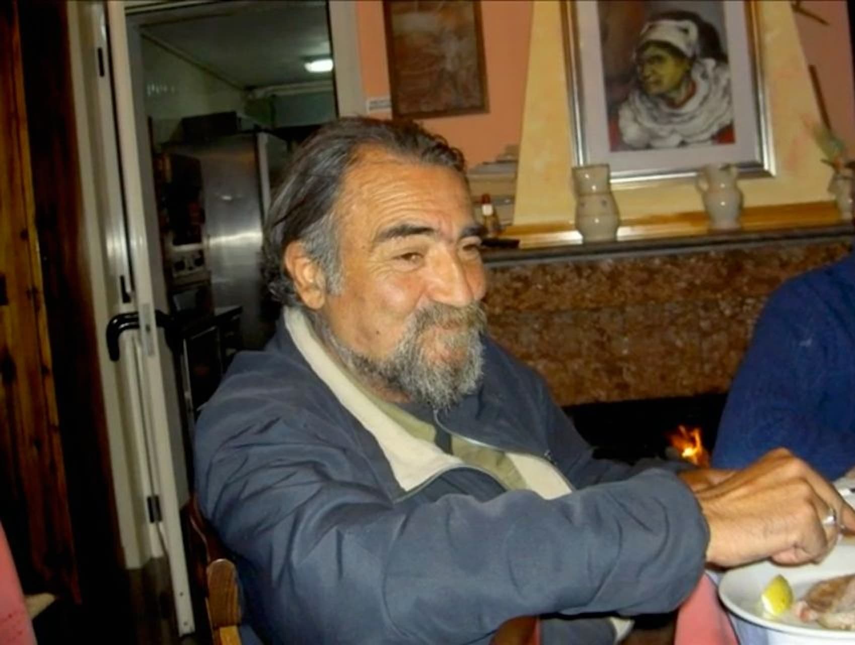 Enzo Schillizzi seduto a tavola, sorridente; alle spalle un ritratto di donna in costume