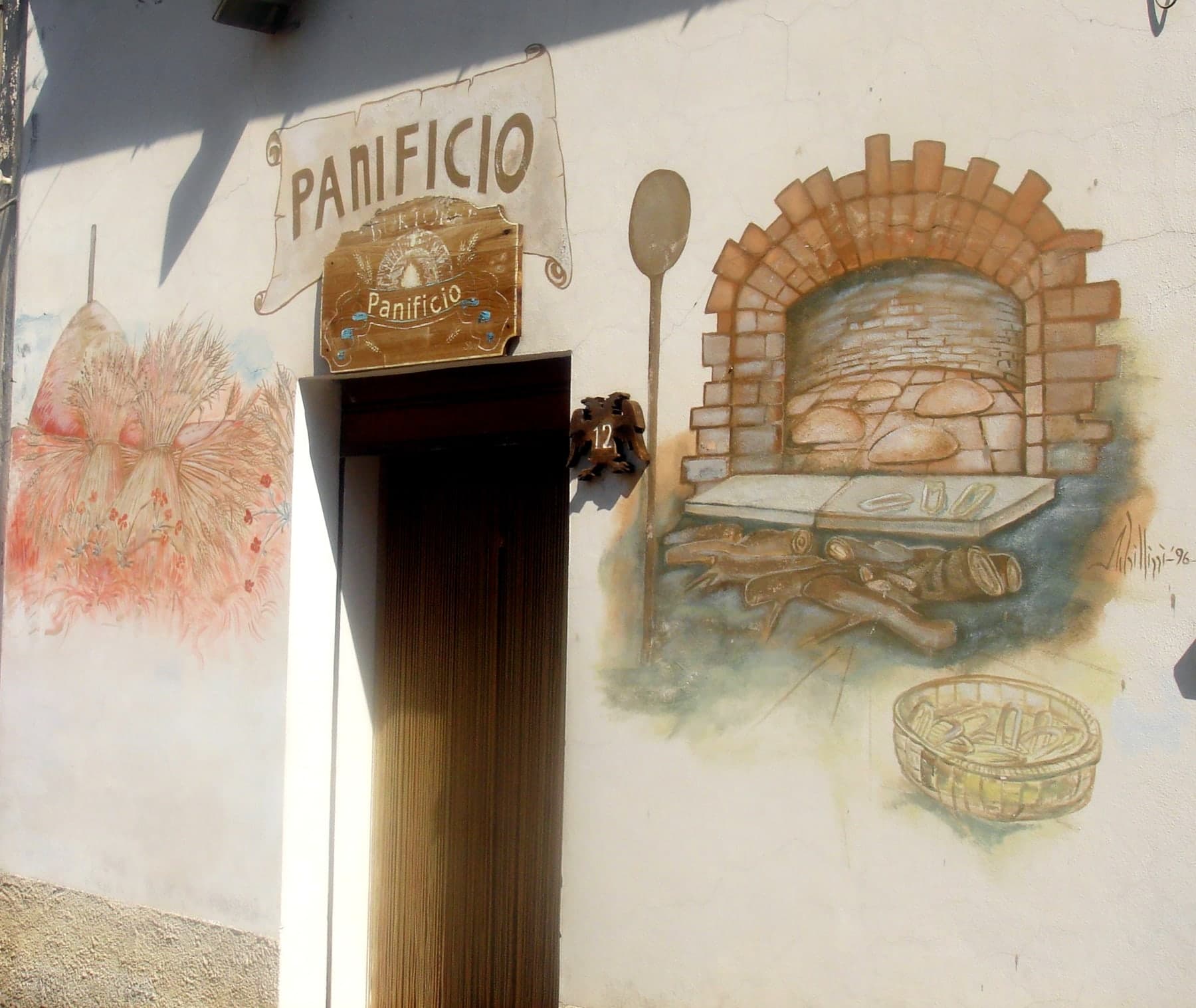 Accanto alla porta del panificio, il forno a legna con il pane, la pala, le fascine e un cesto.