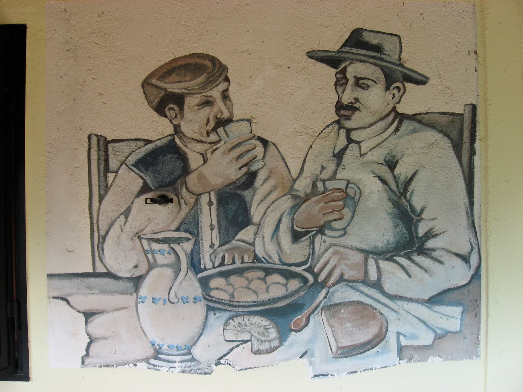 Due uomini con il cappello bevono a tavola: la brocca, il pane, una ciotola sul tavolo. Al ristorante Luna Rossa di Terranova di Pollino.